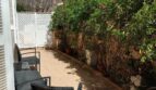 Sonnige Terrasse mit Rattanmöbeln, Fliesenboden und üppigem Grünbewuchs an weißer Gartenmauer des Reihenhauses.