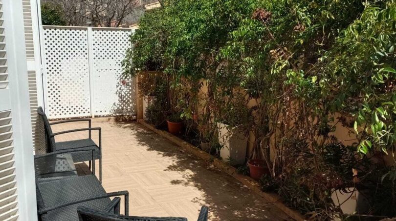 Sunny terrace with rattan furniture, tile floor and lush green vegetation at the white garden wall of the townhouse.