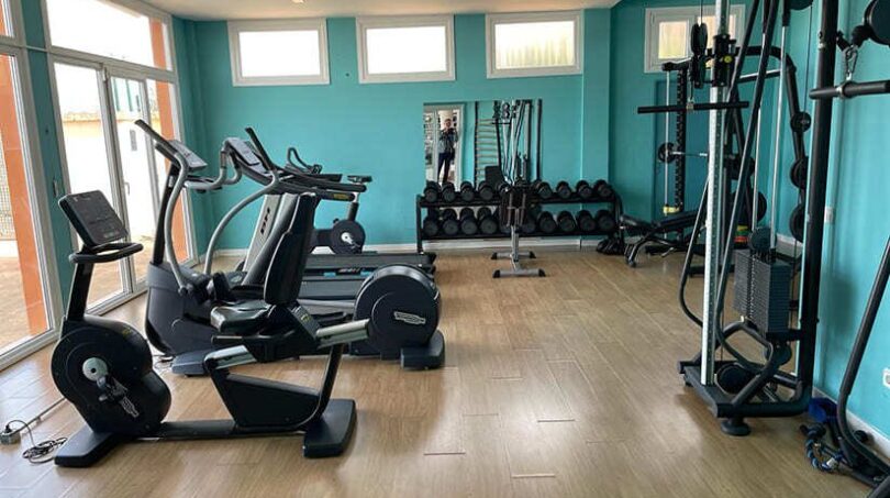 Moderner Fitnessraum mit Cardiogeräten, Hanteln und Cable-Machine, türkisfarbene Wände, Holzboden, Apartment El Toro