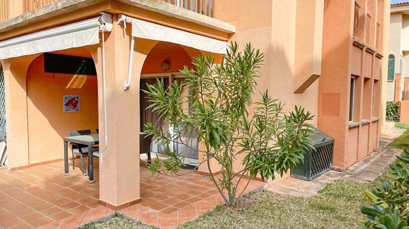 Überdachte Terrasse mit Markise und Gartenmöbeln, umgeben von mediterranem Grün und gepflegtem Rasen.
