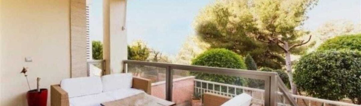 Überdachte Terrasse mit Rattan-Lounge, Glasgeländer und Blick auf mediterrane Bäume, Palma de Mallorca.
