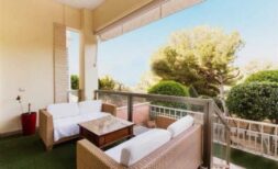 Überdachte Terrasse mit Rattan-Lounge, Glasgeländer und Blick auf mediterrane Bäume, Palma de Mallorca.