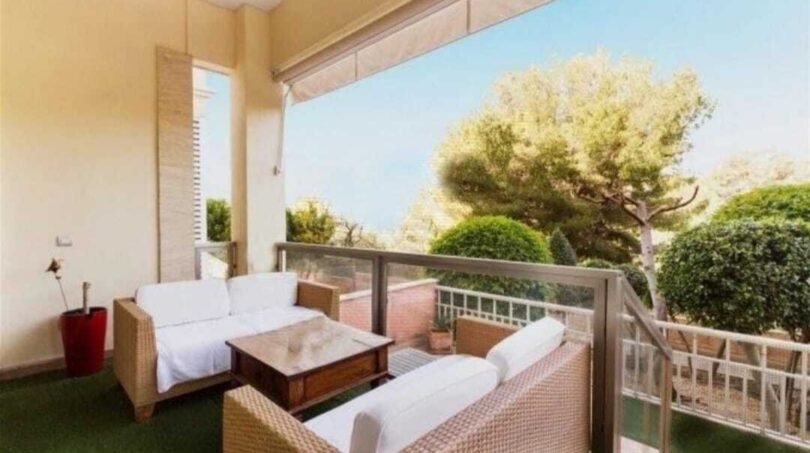 Überdachte Terrasse mit Rattan-Lounge, Glasgeländer und Blick auf mediterrane Bäume, Palma de Mallorca.