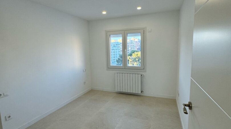 Helles, leeres Schlafzimmer im Penthouse mit weißen Wänden, Einbauspots, Heizkörper und Stadtblick aus dem Fenster.