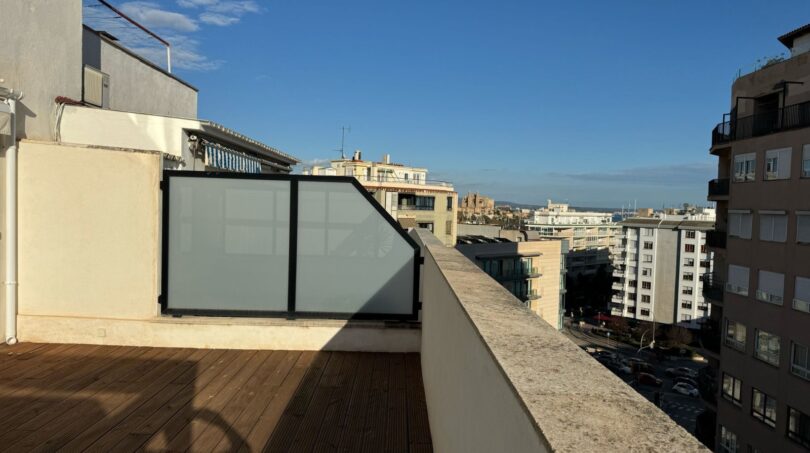 Sonnige Dachterrasse mit Holzboden, Glastrennwand und Panoramablick über Palma de Mallorca bei blauem Himmel.