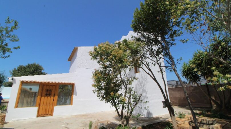 Außenansicht der weißen Finca in Son Ferriol mit Holztür, Terrakottadach und mediterranem Garten bei blauem Himmel.