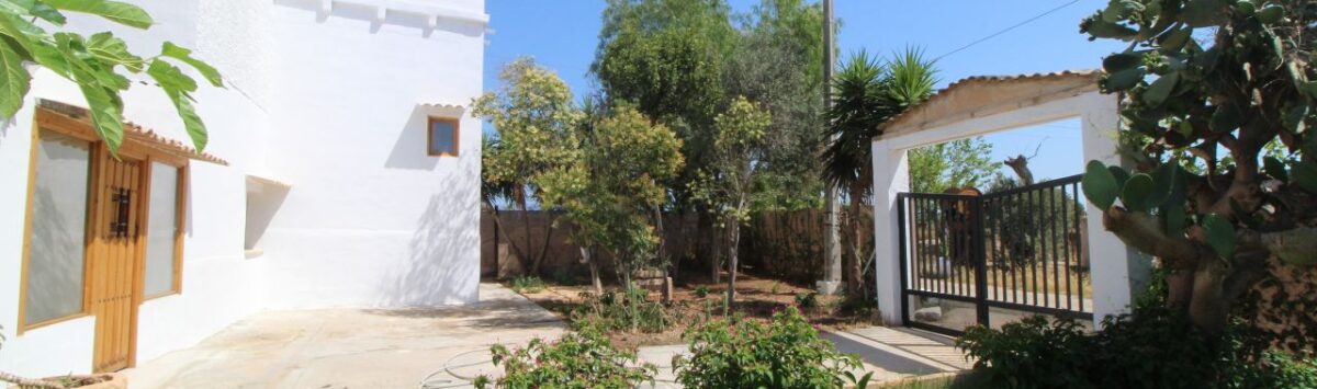 Außenbereich einer Finca in Son Ferriol mit weißer Fassade, Gartenanlage, Einfahrtstor und mediterranen Pflanzen bei blauem Himmel.