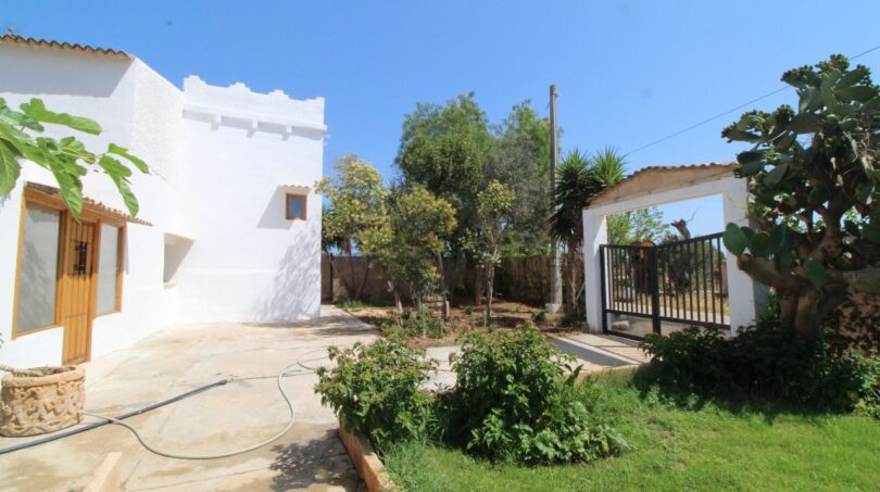 Außenbereich einer Finca in Son Ferriol mit weißer Fassade, Gartenanlage, Einfahrtstor und mediterranen Pflanzen bei blauem Himmel.