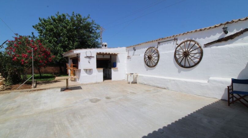 Sonniger Innenhof der Finca mit weißer Hausfassade, Wagenrädern und Blick auf üppige Bepflanzung.