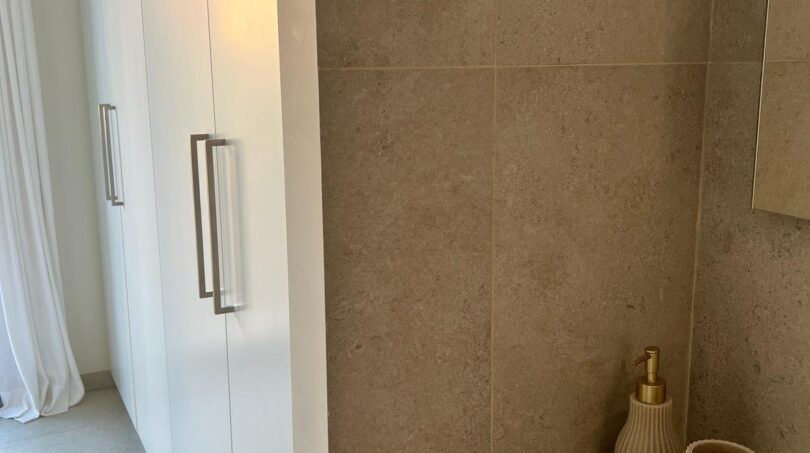 Modernes Luxusbad mit beigen Natursteinwänden, weißen Einbauschränken und eleganten Accessoires in Gold in Palma de Mallorca