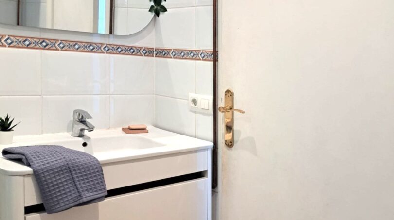 Modernes weißes Badezimmer mit Waschtischunterschrank, Rundspiegel und dekorativer Pflanze in Strandwohnung Palma