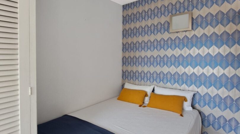 Helles Schlafzimmer mit Doppelbett, blauer Tagesdecke, gelben Kissen und dekorativem Tapetenwandmuster in Strandwohnung Palma.