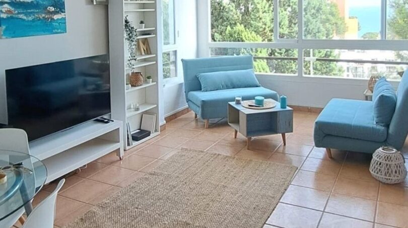 Helles Wohnzimmer mit blauen Möbeln, Flachbild-TV und Panoramafenster mit Meerblick in Palma de Mallorca.