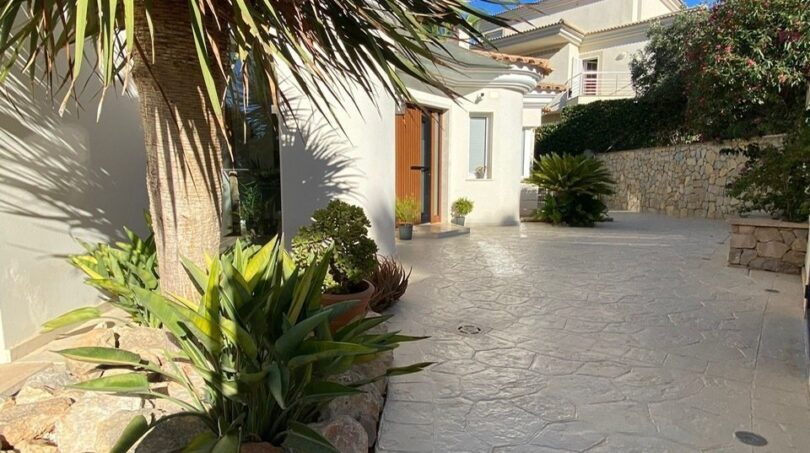 Entrada de una villa moderna en Santa Ponça con palmeras, cantero de piedra y pavimento de hormigón claro bajo cielo azul.