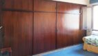 Schlafzimmer mit großem Einbauschrank aus dunklem Holz, blauem Teppichboden und Fenster, renovierungsbedürftig