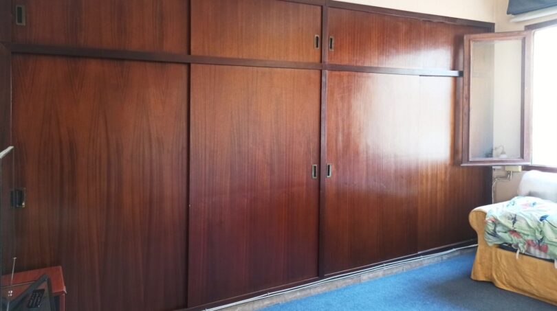 Schlafzimmer mit großem Einbauschrank aus dunklem Holz, blauem Teppichboden und Fenster, renovierungsbedürftig