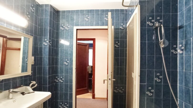 Badezimmer mit blauen Fliesen, Waschbecken, Dusche und Spiegel – renovierungsbedürftig, aber funktional.