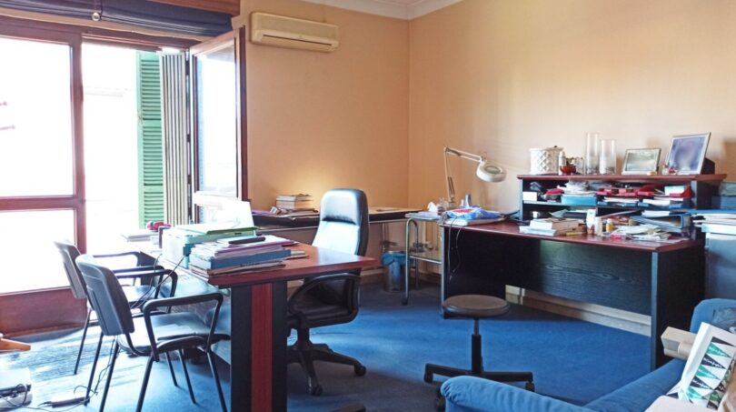 Bürozimmer mit blauem Teppich, zwei Schreibtischen, Klimaanlage und großem Fenster mit Fensterläden.