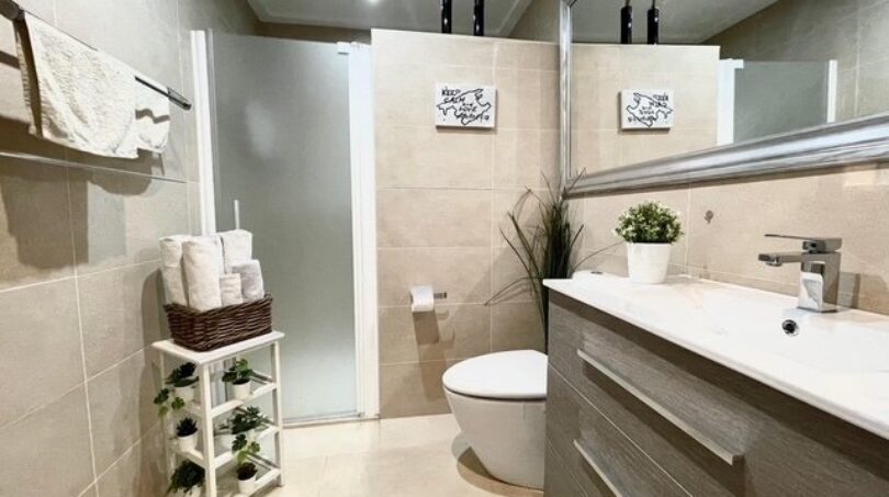 Modernes Badezimmer mit beiger Fliesenverkleidung, Glasdusche, Wandhänge-WC und elegantem Holzwaschtisch in Llucmajor.