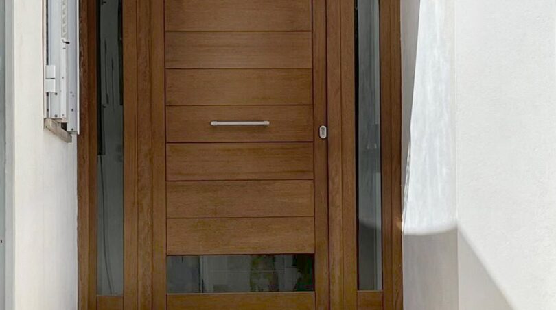 Eingangsbereich des Reihenhauses in Llucmajor mit moderner Holztür, Glasseiten, Designerlampe und Natursteintreppe.