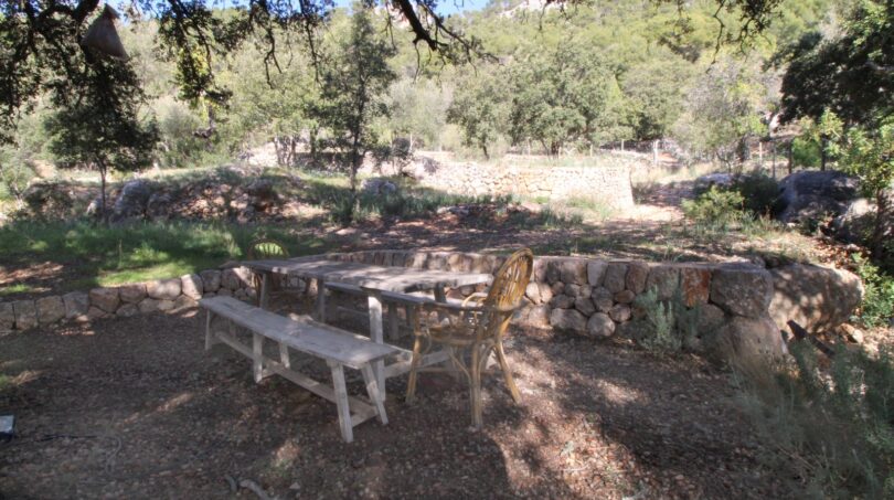 Rustikaler Außensitzbereich mit Holzbank und Rattanstuhl, umgeben von Natursteinmauern und Eichen in Esporles.