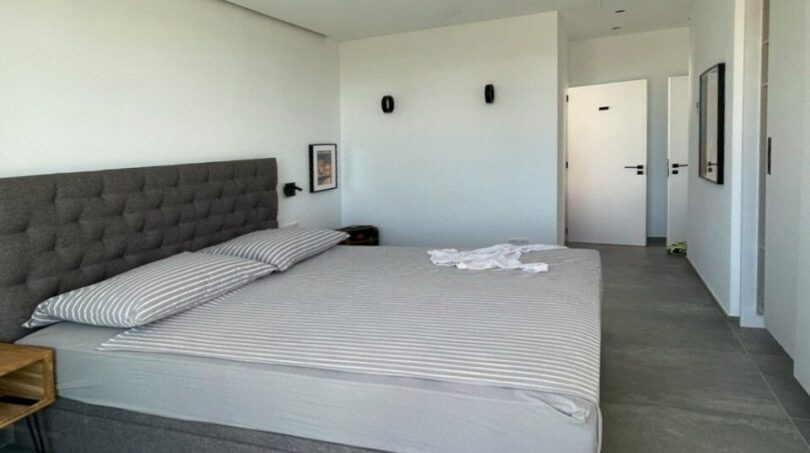 Helles Schlafzimmer mit Doppelbett, gepolstertem Kopfteil, Deckenventilator und großen Bodenfliesen in Meeresnähe.