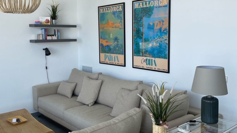 Helles Wohnzimmer mit grauem Sofa, Holzcouchtisch und Mallorca-Vintage-Postern an weißer Wand.