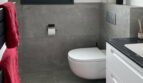 Modernes Badezimmer mit Wandhänge-WC, grauer Betonoptik-Fliesen, weißem Waschtisch und rotem Akzent-Handtuch.