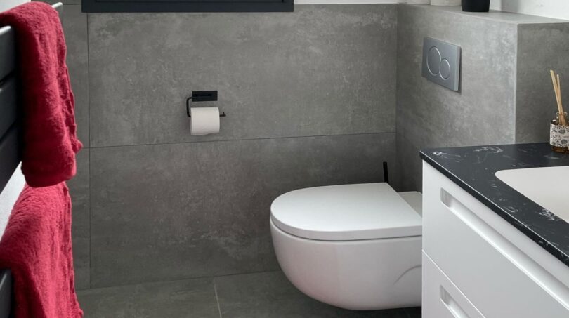 Modernes Badezimmer mit Wandhänge-WC, grauer Betonoptik-Fliesen, weißem Waschtisch und rotem Akzent-Handtuch.