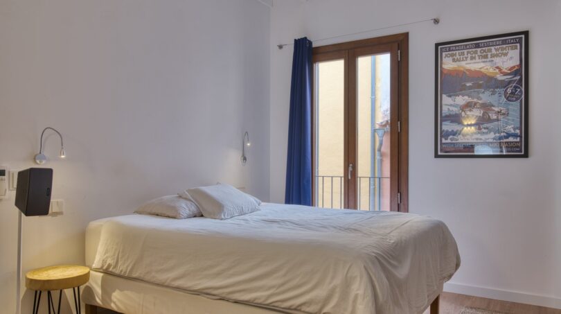 Helles Schlafzimmer mit Doppelbett, Holzbalken, Balkon und blauem Vorhang im Stadthaus Alaró