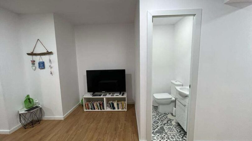Helles Wohnzimmer mit Holzboden, TV-Regal mit Büchern und Blick in modernes Bad mit Zementfliesen in Inca.