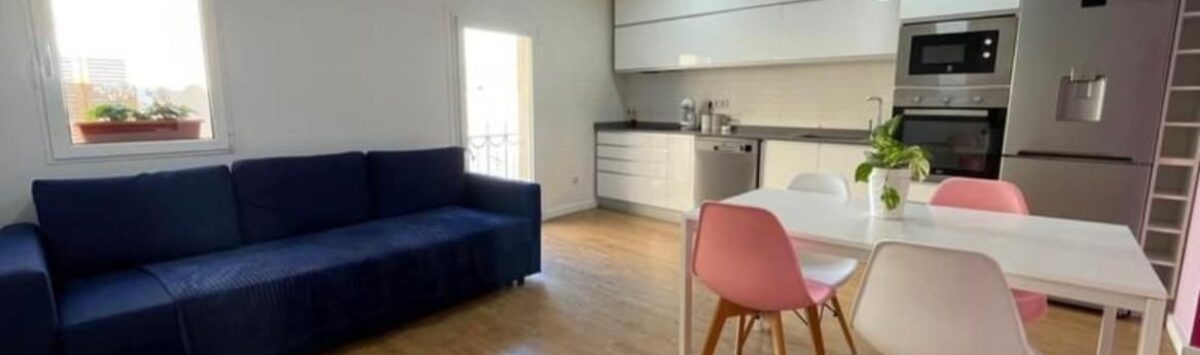 Área abierta de sala-comedor con sofá azul, mesa de comedor blanca, sillas rosas y cocina integrada moderna en Inca.