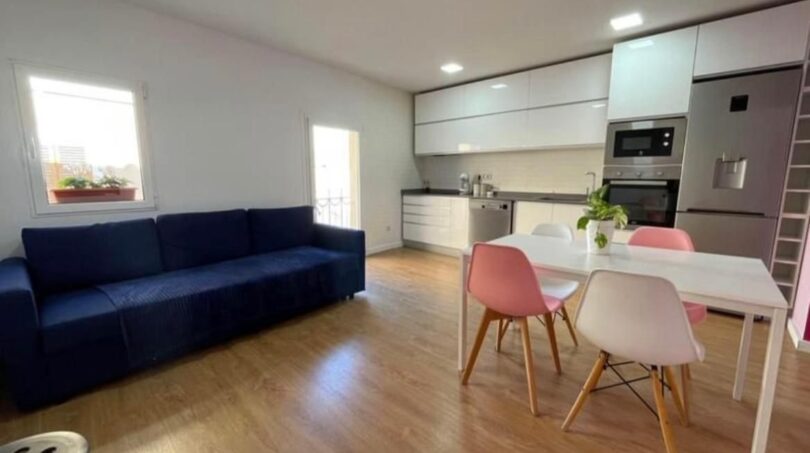 Área abierta de sala-comedor con sofá azul, mesa de comedor blanca, sillas rosas y cocina integrada moderna en Inca.