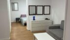 Helles Wohnzimmer mit grauem Sofa, weißem Couchtisch und Blick ins Schlafzimmer, Holzboden, moderne Ausstattung