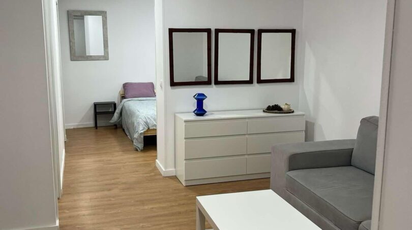 Helles Wohnzimmer mit grauem Sofa, weißem Couchtisch und Blick ins Schlafzimmer, Holzboden, moderne Ausstattung