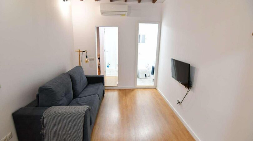 Sala de estar luminosa con sofá gris, suelo de madera, aire acondicionado y vigas de madera en Inca