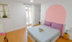 Helles Schlafzimmer mit Doppelbett, lila Bettwäsche, rosa Wandbogen und Holzparkett in Inca