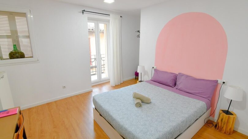Helles Schlafzimmer mit Doppelbett, lila Bettwäsche, rosa Wandbogen und Holzparkett in Inca