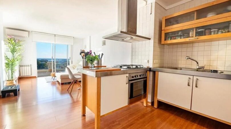 Offene Küche mit Kochinsel, Edelstahlabzug und Holzboden, Blick auf lichtdurchfluteten Wohnbereich mit Meerblick-Terrasse