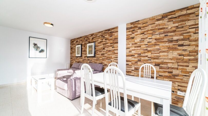 Heller Wohn-Essbereich mit weißem Tisch, lila Sofa und rustikaler Steinwand in exklusiver Wohnung in Artà.