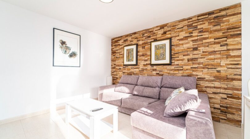 Wohnzimmer der Wohnung in Artà mit grauem Ecksofa, Holzsteinwand und modernen Kunstwerken