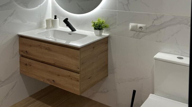 Modernes Badezimmer mit rundem LED-Spiegel, schwebendem Holzwaschtisch und Marmorfliesen in sanierter Wohnung Palma