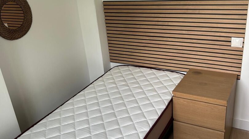 Kleines Schlafzimmer mit Einzelbett, Holz-Lattenoptik-Kopfteil, Nachttisch und Rattanspiegeln an weißer Wand.