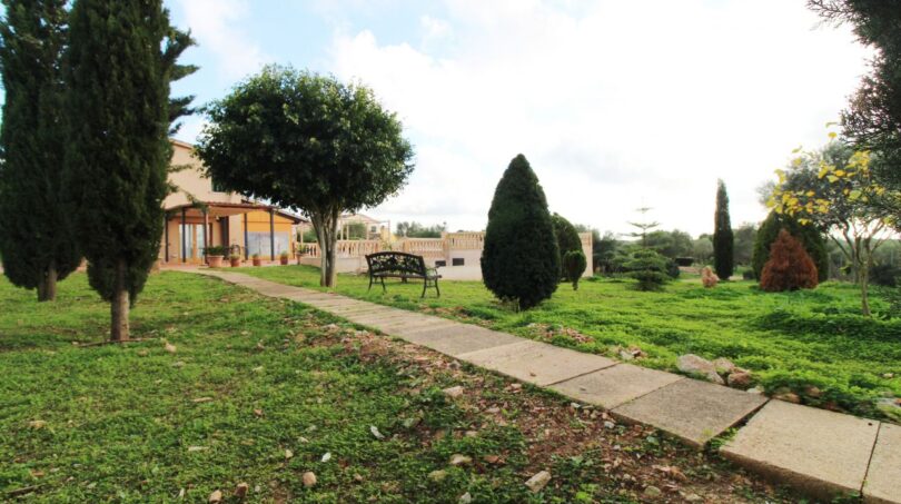 Weitläufiger Garten der Finca mit gepflegten Zypressen, Steinweg, Gartenbank und Blick auf das Haupthaus.
