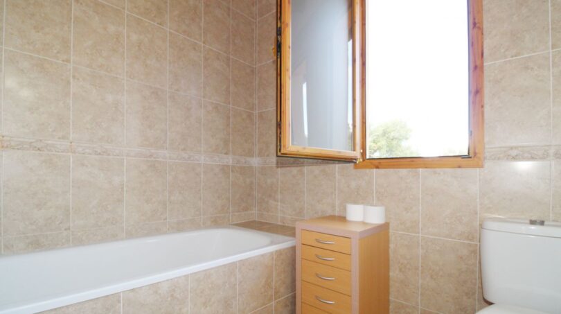 Badezimmer der Finca mit Badewanne, Holzfenster, beigen Fliesen und kleinem Rollcontainer