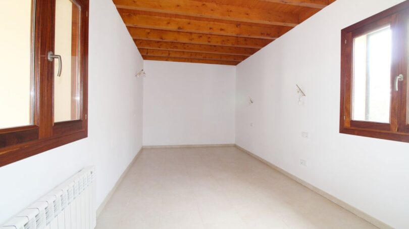 Leeres Zimmer mit Holzbalkendecke, Fliesenboden, Heizkörper und zwei Holzfenstern in Neubau-Finca Palma