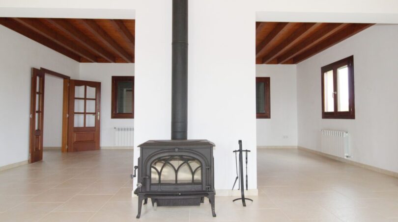 Geräumiges Wohnzimmer der Neubau-Finca mit Holzofen, Holzbalkendecke und hellem Fliesenboden.
