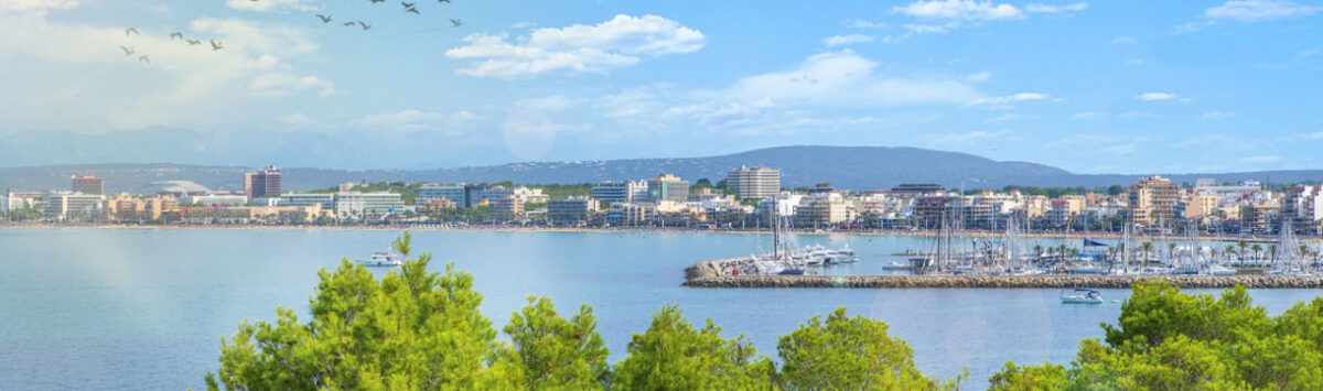 Vista panorámica de la costa de Llucmajor con puerto deportivo, mar turquesa y silueta de la ciudad ante montañas.