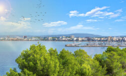 Panoramic view of Llucmajor's coast with marina, turquoise sea and city skyline in front of mountains.