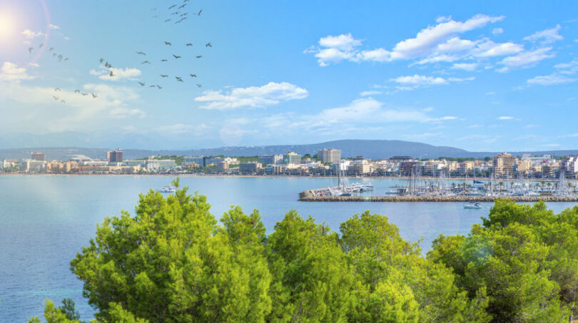Vista panorámica de la costa de Llucmajor con puerto deportivo, mar turquesa y silueta de la ciudad ante montañas.